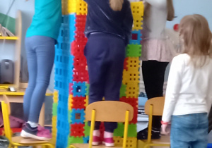 Cztery dziewczynki stoją na krzesełkach budują wysoką wieże z kolorowych